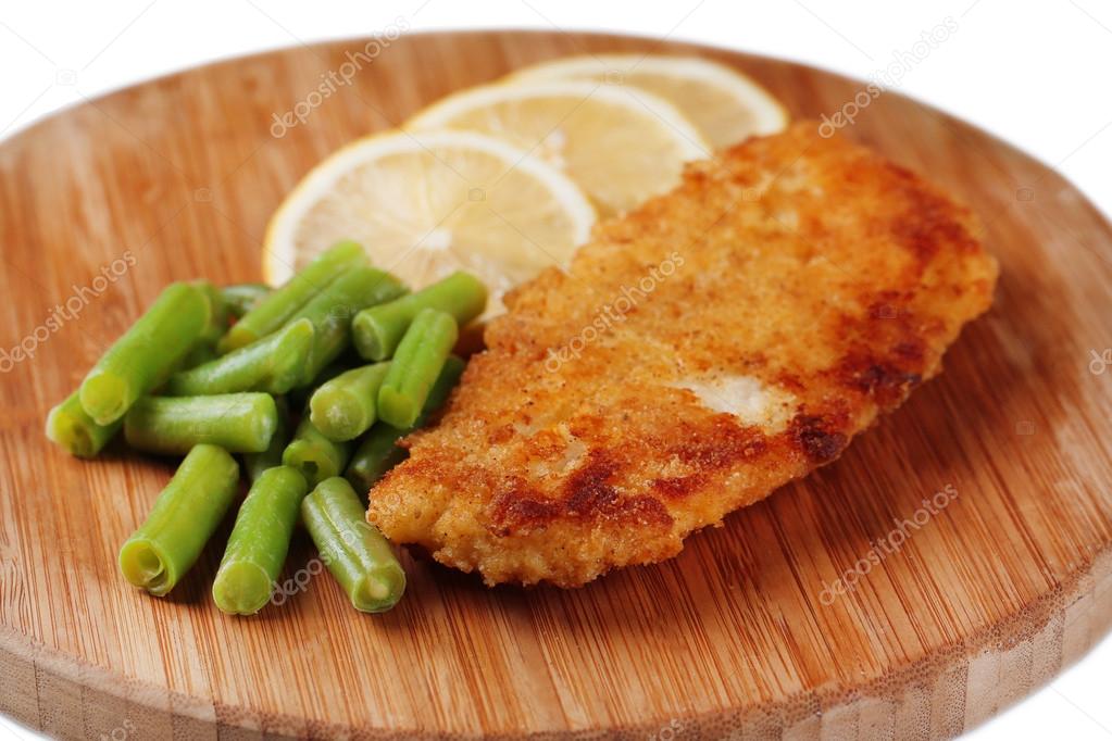 Breaded fried fish fillet and potatoes with asparagus and sliced lemon on wooden cutting board isolated on white