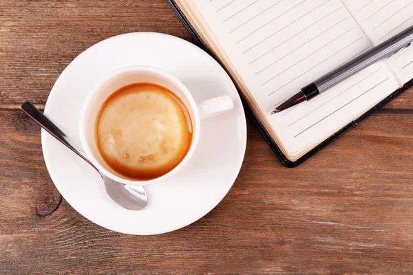 Tasse Kaffee auf Untertasse mit Tagebuch, Stift und Löffel auf Holztischhintergrund — Stockfoto