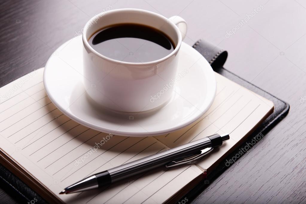 Cup of coffee on saucer with diary and pen on wooden table background