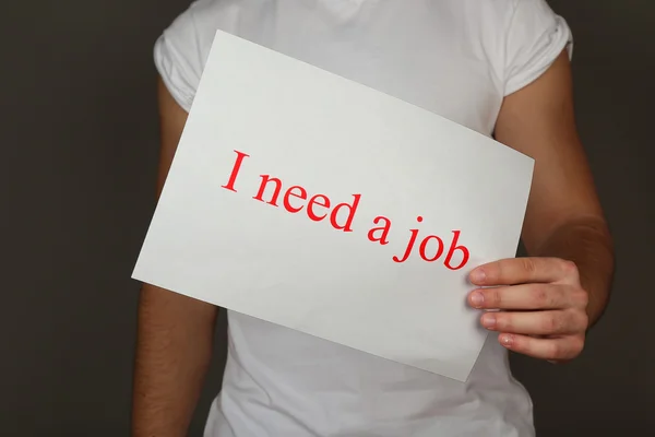 Foglio di carta con iscrizione Ho bisogno di un lavoro in mano maschile su sfondo scuro — Foto Stock