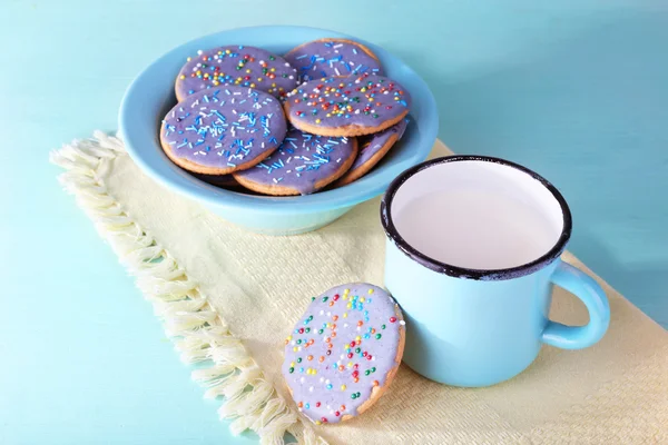 Plate of glazed cookies and mug of milk on napkin and color wooden table background — Stock Photo, Image