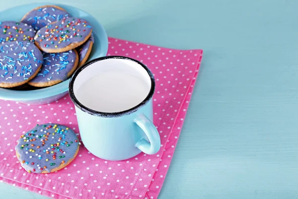 Plate of glazed cookies and mug of milk on napkin and color wooden table background — Stock Photo, Image