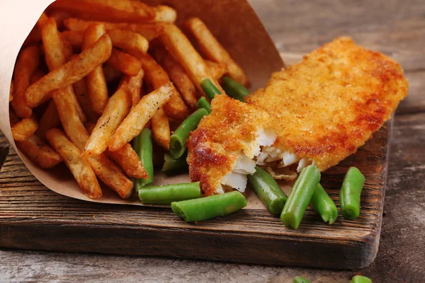 Breaded fried fish fillet and potatoes in paper bag with asparagus on cutting board and rustic wooden background — Stock Photo, Image