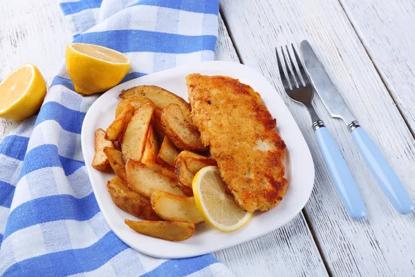 Filetto di pesce fritto impanato e patate con limone affettato sul piatto con tovagliolo su sfondo di tavole di legno di colore — Foto Stock