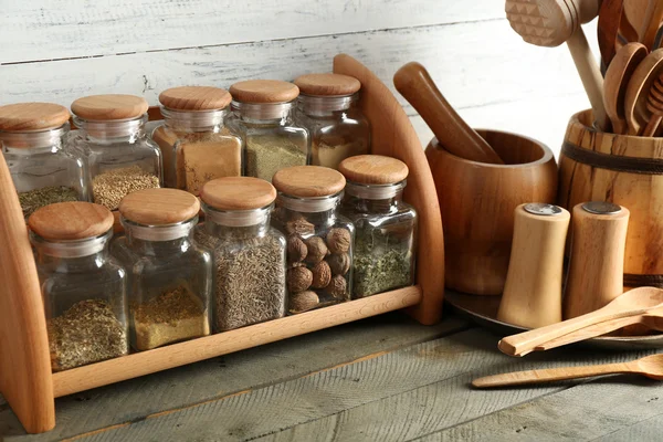 Verschillende kruiden in glazen flessen op stand met gebruiksvoorwerpen op houten tafel en planken achtergrond — Stockfoto