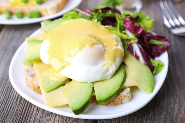 Roti panggang dengan telur Benedict dan alpukat di piring di atas meja kayu — Stok Foto