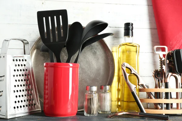 Plastic kitchen utensils in red cup with bottle of olive oil on wooden background — Stock Photo, Image