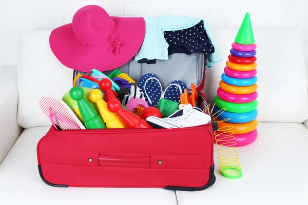 Suitcase packed with clothes and child toys on white sofa and brick wall background — Stock Photo, Image