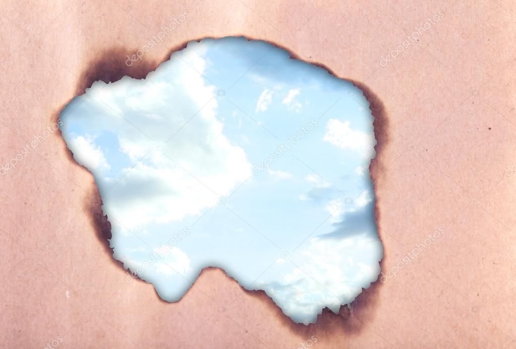 Cloudy sky through scorched hole in paper