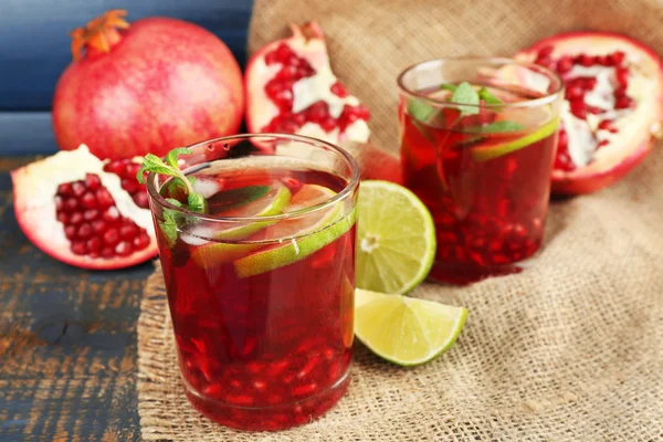 Pomegranate drink in glasses with slices of lime and mint on burlap cloth and color wooden background — Stock Photo, Image