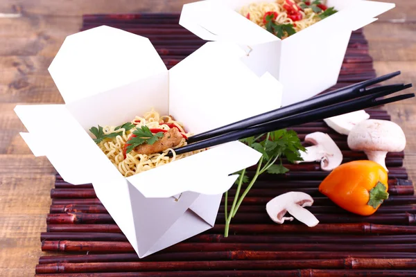 Chinese noodles in takeaway boxes on bamboo mat on wooden background — Stock Photo, Image