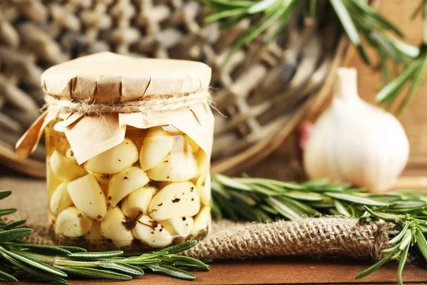 Ingeblikte knoflook in glazen pot en rieten mat en rozemarijn takken, op houten achtergrond — Stockfoto