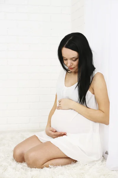 Nuori raskaana oleva nainen istuu matolla kevyellä taustalla — kuvapankkivalokuva