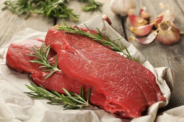 Raw beef steak with rosemary and garlic on paper on wooden background — Stock Photo, Image