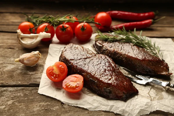 Composition avec une délicieuse viande rôtie sur feuille de papier, tomates et brins de romarin sur fond bois — Photo