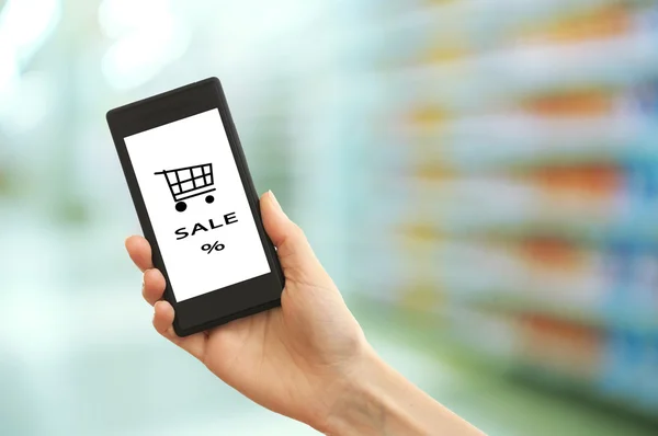 Mobiele winkelen concept. Hand met mobiele telefoon voor internet winkelen op supermarkt achtergrond — Stockfoto