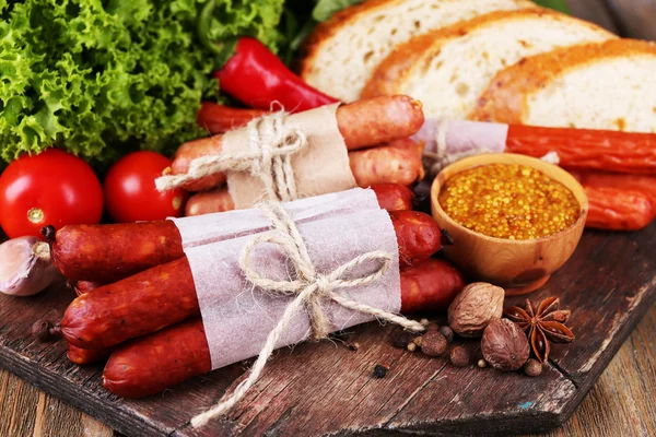 Assortment of thin sausages, bread, mustard in bowl and spices on cutting board, on wooden background — Stock Photo, Image