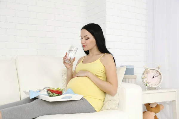 Jovem grávida relaxante no sofá com vidro de água e salada em casa interior fundo — Fotografia de Stock