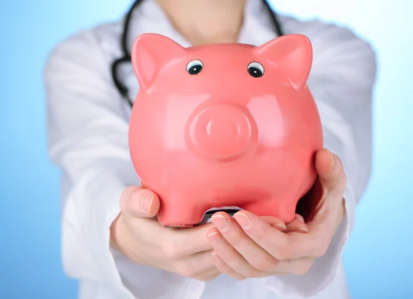 Doutor com o banco piggy rosa no fundo azul, close-up — Fotografia de Stock