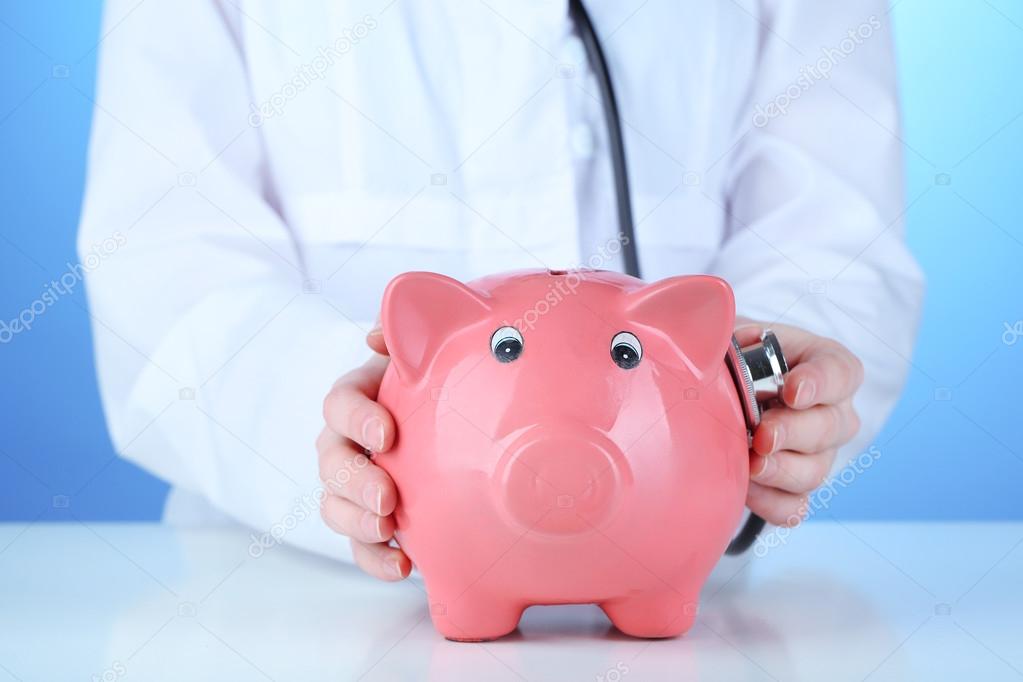 Doctor with pink piggy bank on blue background, close up