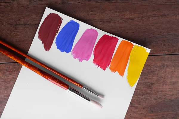 Colpi di vernice colorata con pennello su foglio bianco di carta su sfondo tavolo di legno — Foto Stock