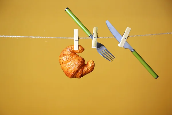 Fork, knife and croissant hanging from clothesline on color background — Stock Photo, Image