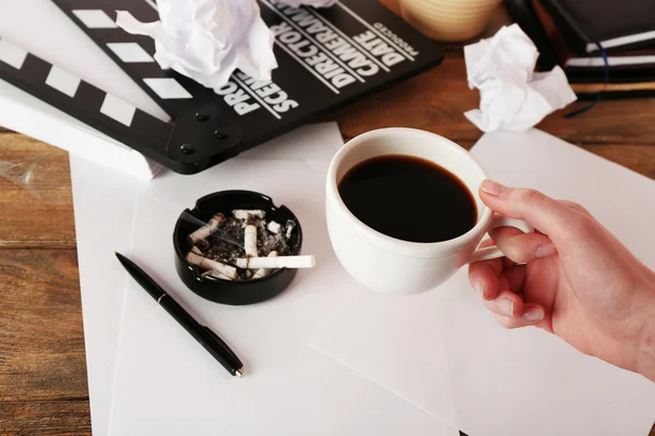 Female hand with cup of coffee, moving clapper and sheets of paper on wooden background — Stock Photo, Image