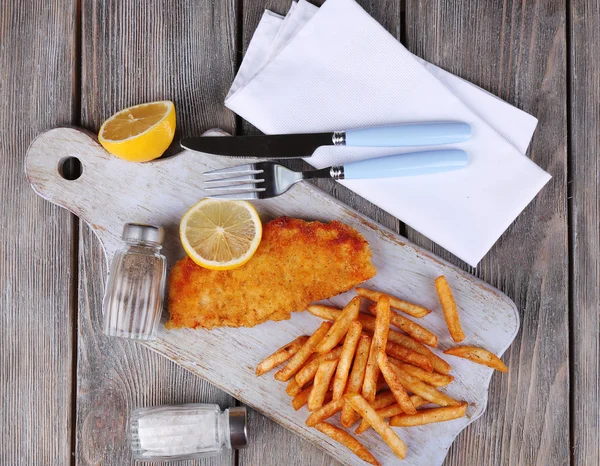 Breaded fried fish fillet and potatoes with sliced lemon and cutlery on cutting board and wooden planks background — Stock Photo, Image