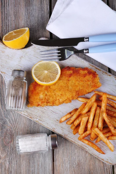 Breaded fried fish fillet and potatoes with sliced lemon and cutlery on cutting board and wooden planks background — Stock Photo, Image