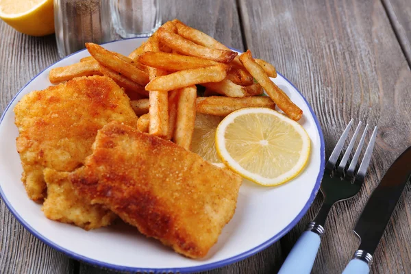 Filetes de pescado frito y patatas con rodajas de limón y cubiertos en el plato y tablones de madera de fondo — Foto de Stock