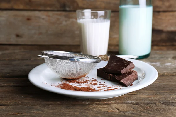 Glas und Flasche Milch mit Schokoladenstücken auf Plat mit Kakaosieb auf rustikalem Holzplanken Hintergrund — Stockfoto