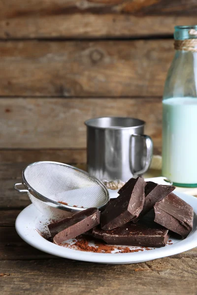 Metal mug and glass bottle of milk with chocolate chunks and strainer of cocoa on plate and rustic wooden planks background — Stock Photo, Image