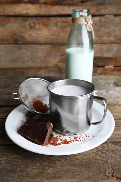 Kovové hrnek a skleněná láhev mléka s kousky čokolády a sítko kakaem na desku a rustikální Dřevěná prkna zázemí — Stock fotografie