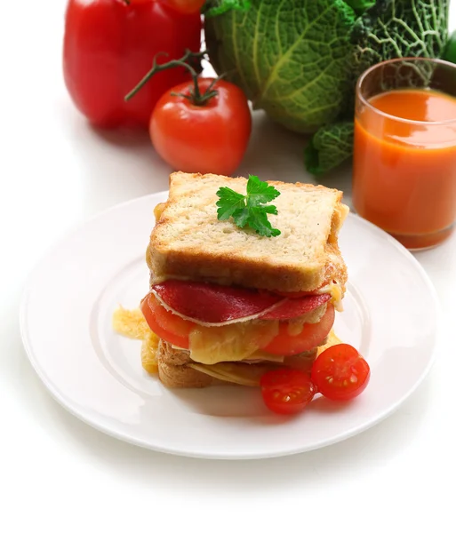 Большой сэндвич с сыром и овощами на тарелке возле стакана сока на свежих овощах на заднем плане — стоковое фото