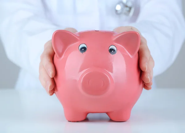 Dokter dengan piggy bank merah muda dengan latar belakang abu-abu, tutup — Stok Foto