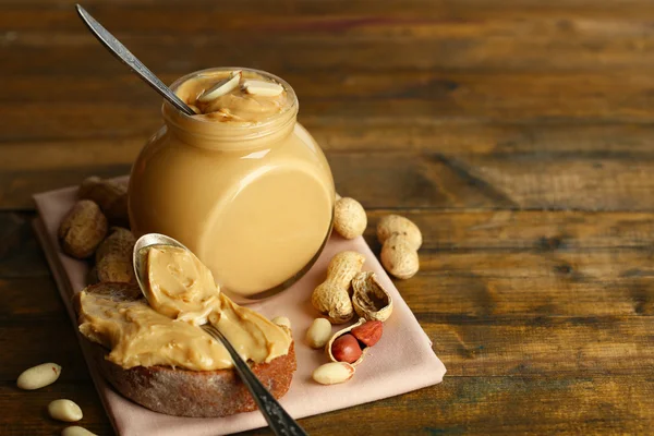 Sabroso sándwich y frasco con mantequilla de maní fresca sobre fondo de madera — Foto de Stock