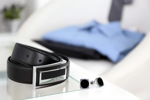 Men's belt and cufflinks on coffee tablewith shirt and tie on chair on background