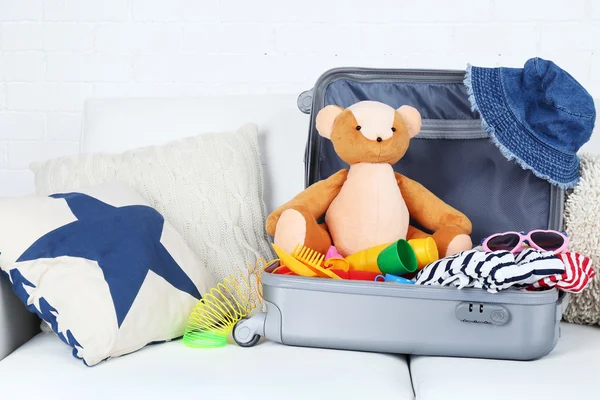 Suitcase packed with clothes and child toys on sofa with pillows and white brick wall background — Stock Photo, Image