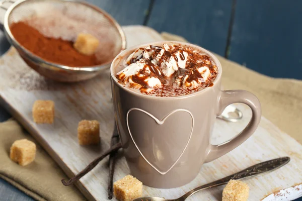 Cup of hot coffee with marshmallow on cutting board with sieve of cocoa, lump sugar and sticks of vanilla on color wooden table background — Stock Photo, Image