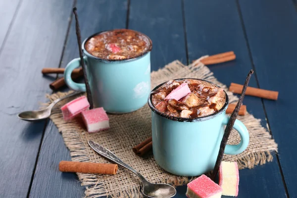 Canecas de café quente com marshmallow em pano de serapilheira com canela e baunilha varas na cor de fundo de mesa de madeira — Fotografia de Stock