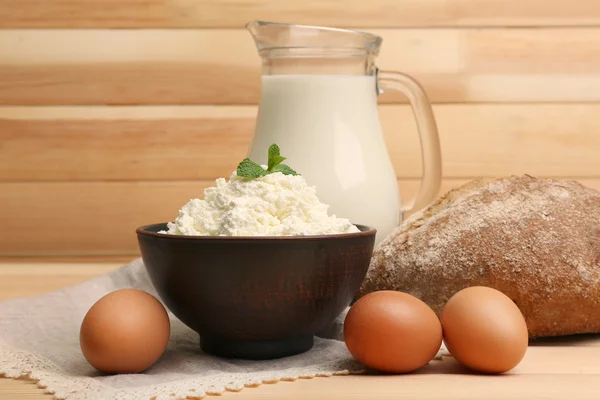 Коттеджный сыр в глиняной миске с кувшином молока, яйцами и буханкой хлеба на фоне деревянных досок — стоковое фото