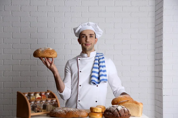 Padeiro na cozinha à mesa com pães frescos no fundo da parede de tijolo branco — Fotografia de Stock