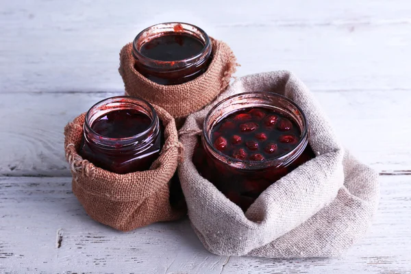 Homemade jars of fruits jam in burlap pouches on color wooden planks background — Stock Photo, Image