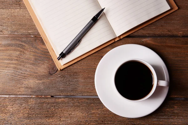 Cup of coffee on saucer with notebook and pen on wooden table background — Stock Photo, Image