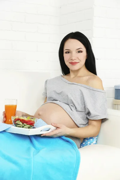 Wanita hamil muda duduk di sofa dengan salad di latar belakang interior rumah — Stok Foto