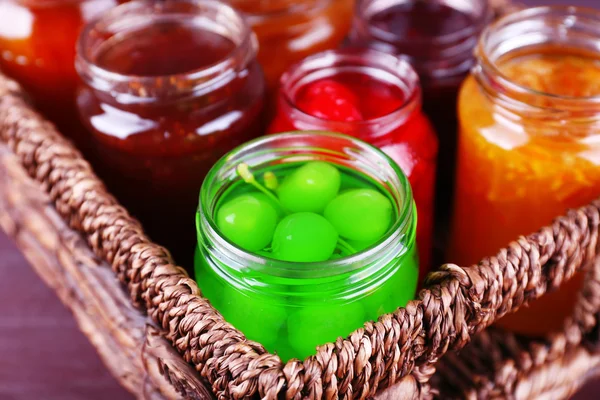 Tarros caseros de mermelada de frutas en canasta de mimbre y fondo de madera de color — Foto de Stock