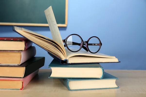 Στοίβα από βιβλία με γυαλιά σε ξύλινο γραφείο, σε πολύχρωμο τοίχο και φόντο μαυροπίνακα — Φωτογραφία Αρχείου
