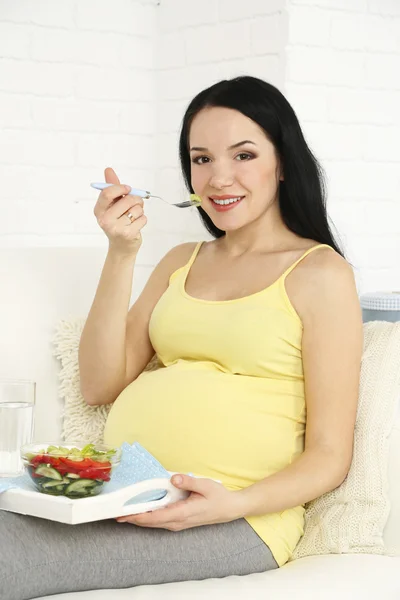 Молода вагітна жінка сидить на дивані з салатом на домашньому внутрішньому тлі — стокове фото