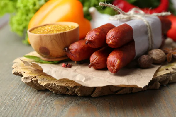 Samenstelling van gerookte dunne worst, mosterd in kom en specerijen op snijplank, op houten achtergrond — Stockfoto