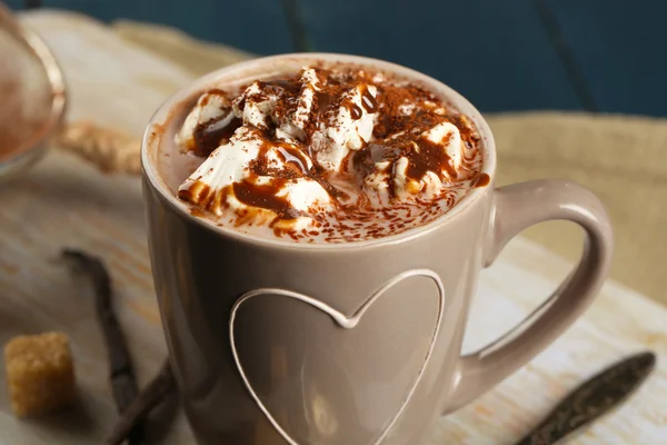 Tasse heißen Kaffee mit Marshmallow auf Schneidebrett mit Sieb aus Kakao, Klumpen Zucker und Stangen Vanille auf farbigem Holztischhintergrund — Stockfoto